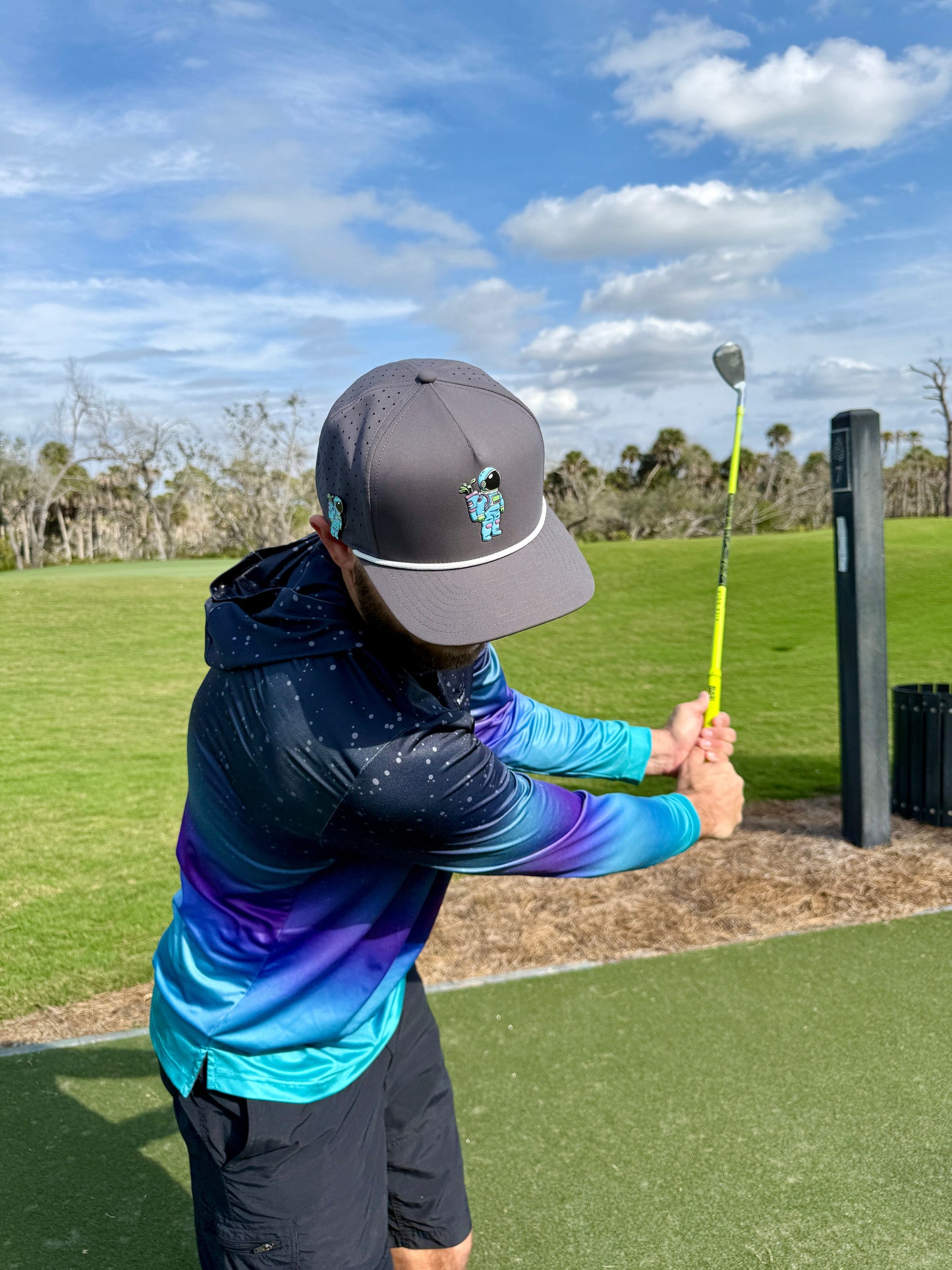 Wearing the gray Astronaut Walker Golf Hat by Logie Joe's Golf, featuring a built-in magnetic ball marker holder, a golfer swings on a grassy course beneath partly cloudy skies, with trees and green landscape in the background.