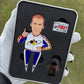 A gloved hand holds a metal case from Pin Creatures with a Ricky Bobby Divot Tool, an enamel pin of a man in a racing suit raising one finger, a small "IF YOU AIN'T FIRST, YOU'RE LAST" sign, and a black clip inside.