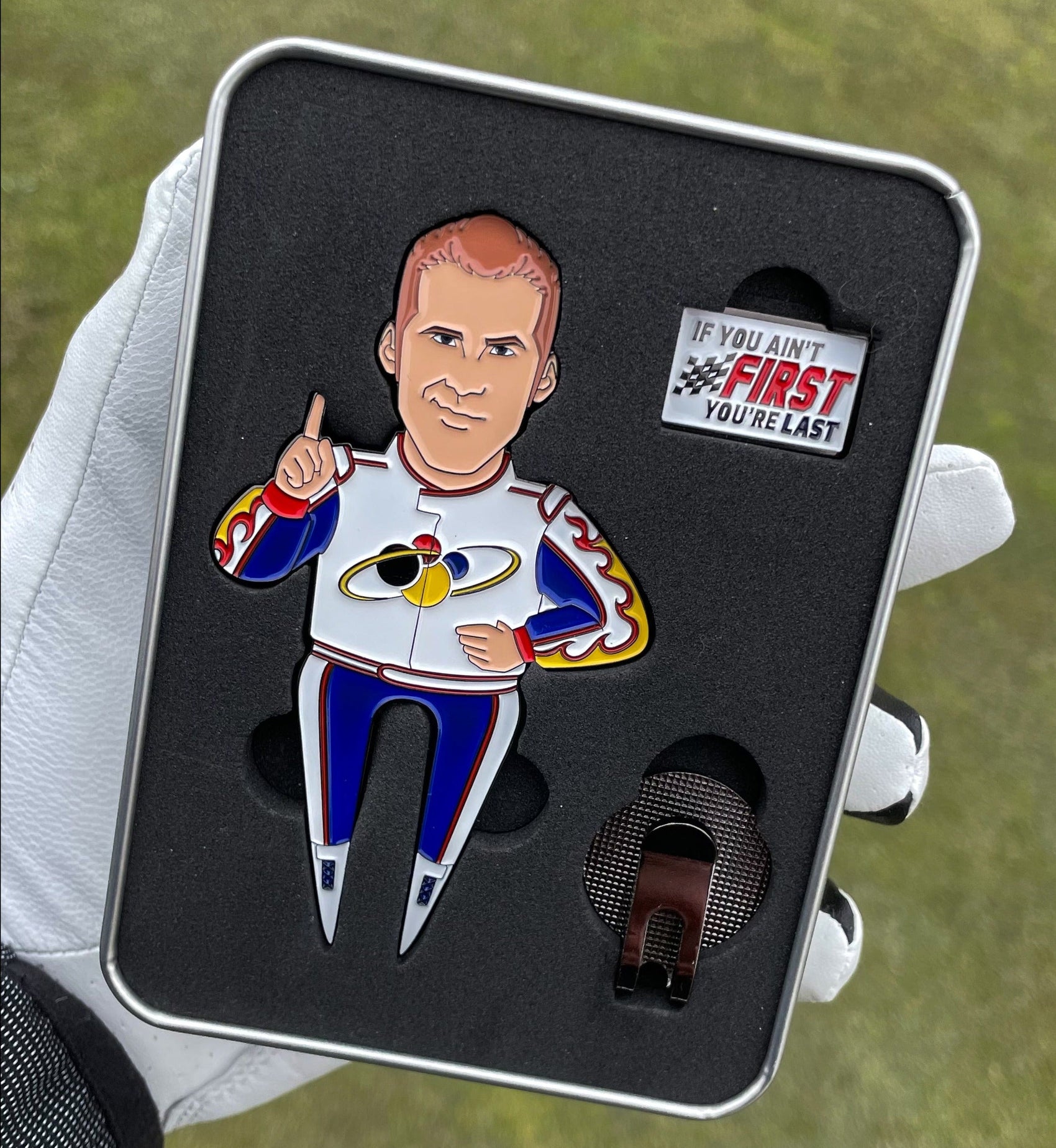 A gloved hand holds a metal case from Pin Creatures with a Ricky Bobby Divot Tool, an enamel pin of a man in a racing suit raising one finger, a small "IF YOU AIN'T FIRST, YOU'RE LAST" sign, and a black clip inside.