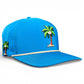 The Logie Joe’s Golf Fairway Palms Golf Hat is a bright blue snapback cap with cartoon palm tree and coconut graphics, a beige cord on the brim, ventilation holes, and an integrated magnetic ball marker holder with marker included. Plain white background.