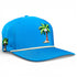 The Logie Joe’s Golf Fairway Palms Golf Hat is a bright blue snapback cap with cartoon palm tree and coconut graphics, a beige cord on the brim, ventilation holes, and an integrated magnetic ball marker holder with marker included. Plain white background.