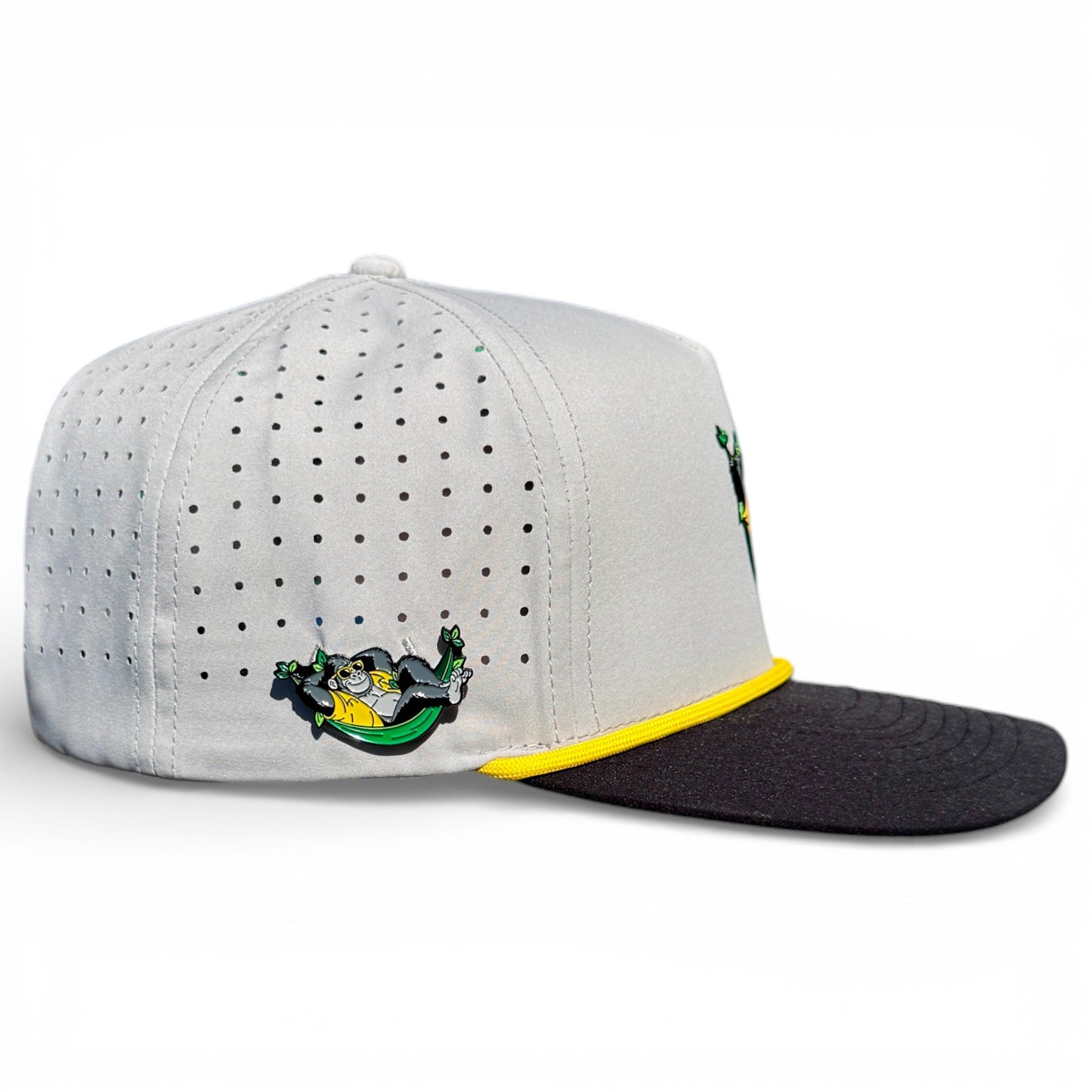 The Banana Hammock Golf Hat by Logie Joe's Golf is a light gray snapback cap with a black brim, yellow trim, perforated side panels, and a lounging cartoon animal in sunglasses. It includes a magnetic ball marker holder and is one-size adjustable.