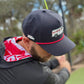 A golfer wears Logie Joe's Golf "Free Golf Balls Van" Snapback Hat with matching ball marker and a black hoodie with red and white hood, swinging a club on a green course. Blurred trees and plants are in the background.