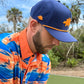 A man stands on a golf course wearing Logie Joe's Golf Dino Nugget Snapback Hat + Matching Ball Marker, featuring navy blue with orange trim, and a bright blue, orange, and white patterned polo shirt; trees and sky in the background.