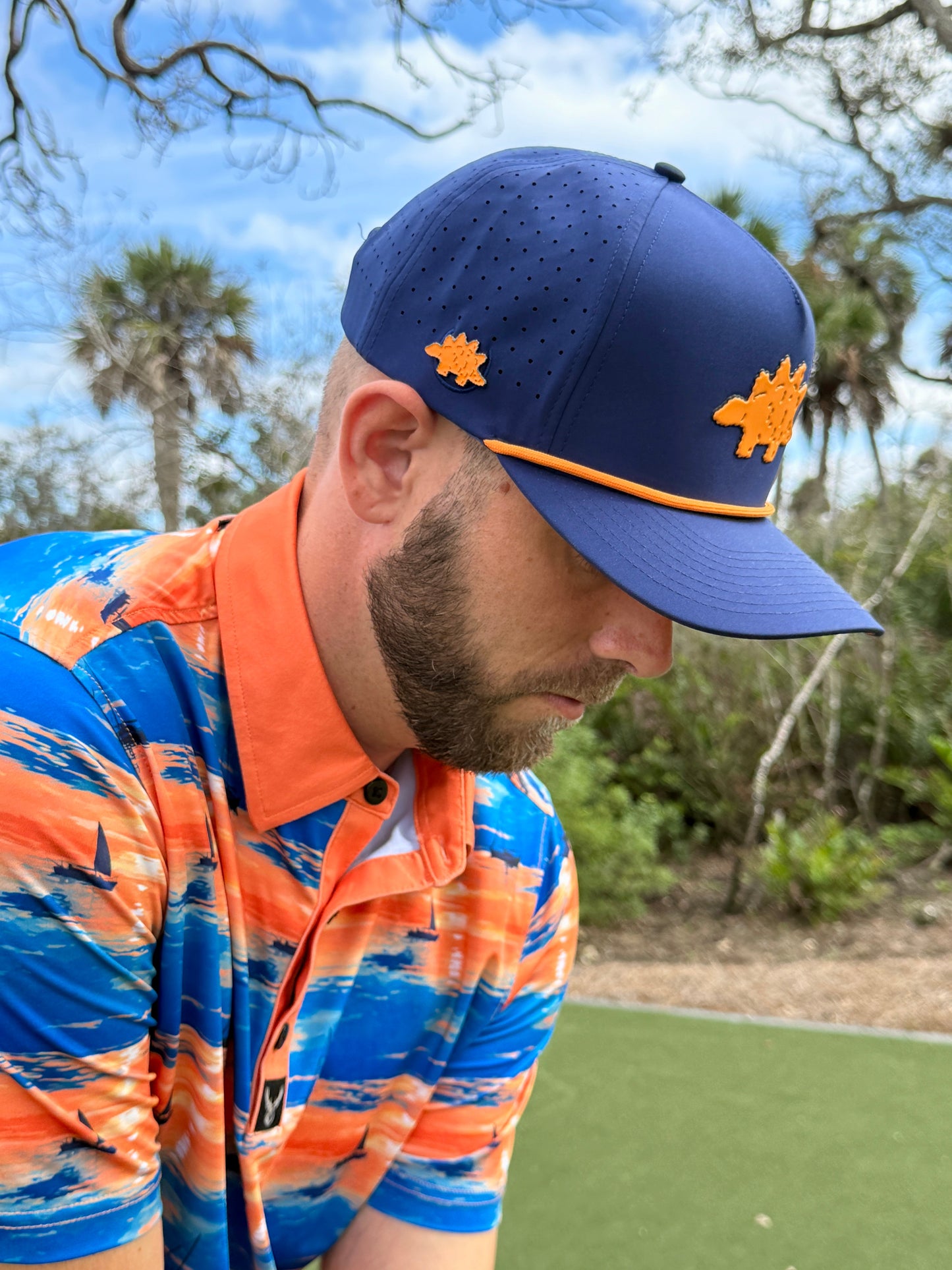 A man stands on a golf course wearing Logie Joe's Golf Dino Nugget Snapback Hat + Matching Ball Marker, featuring navy blue with orange trim, and a bright blue, orange, and white patterned polo shirt; trees and sky in the background.