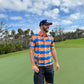 A man wearing a colorful orange and blue shirt, navy shorts, and the Logie Joe's Golf Dino Nugget Snapback Hat with matching ball marker stands on a golf course, holding a club with trees and a partly cloudy sky in the background.