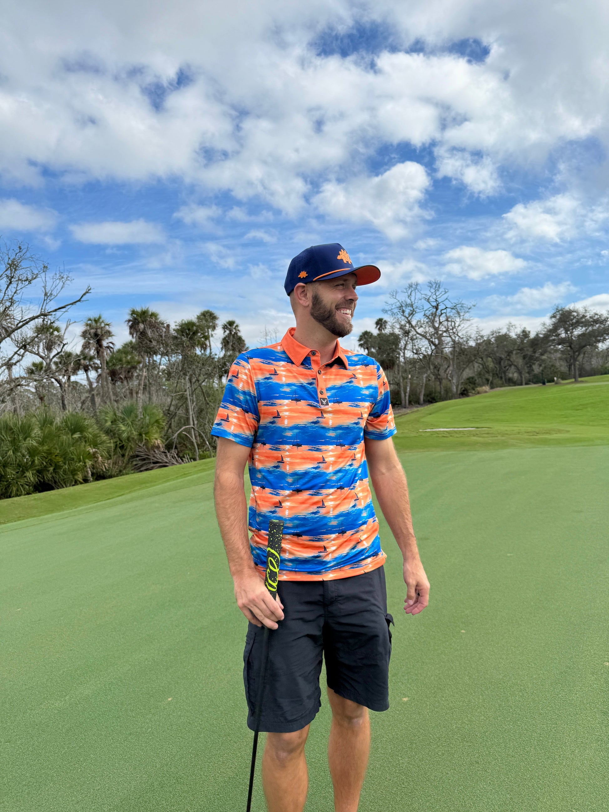 A man wearing a colorful orange and blue shirt, navy shorts, and the Logie Joe's Golf Dino Nugget Snapback Hat with matching ball marker stands on a golf course, holding a club with trees and a partly cloudy sky in the background.