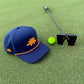 A Logie Joe's Golf Dino Nugget Snapback Hat with a matching ball marker, along with a bright green golf ball and black putter, sit on a well-manicured green.