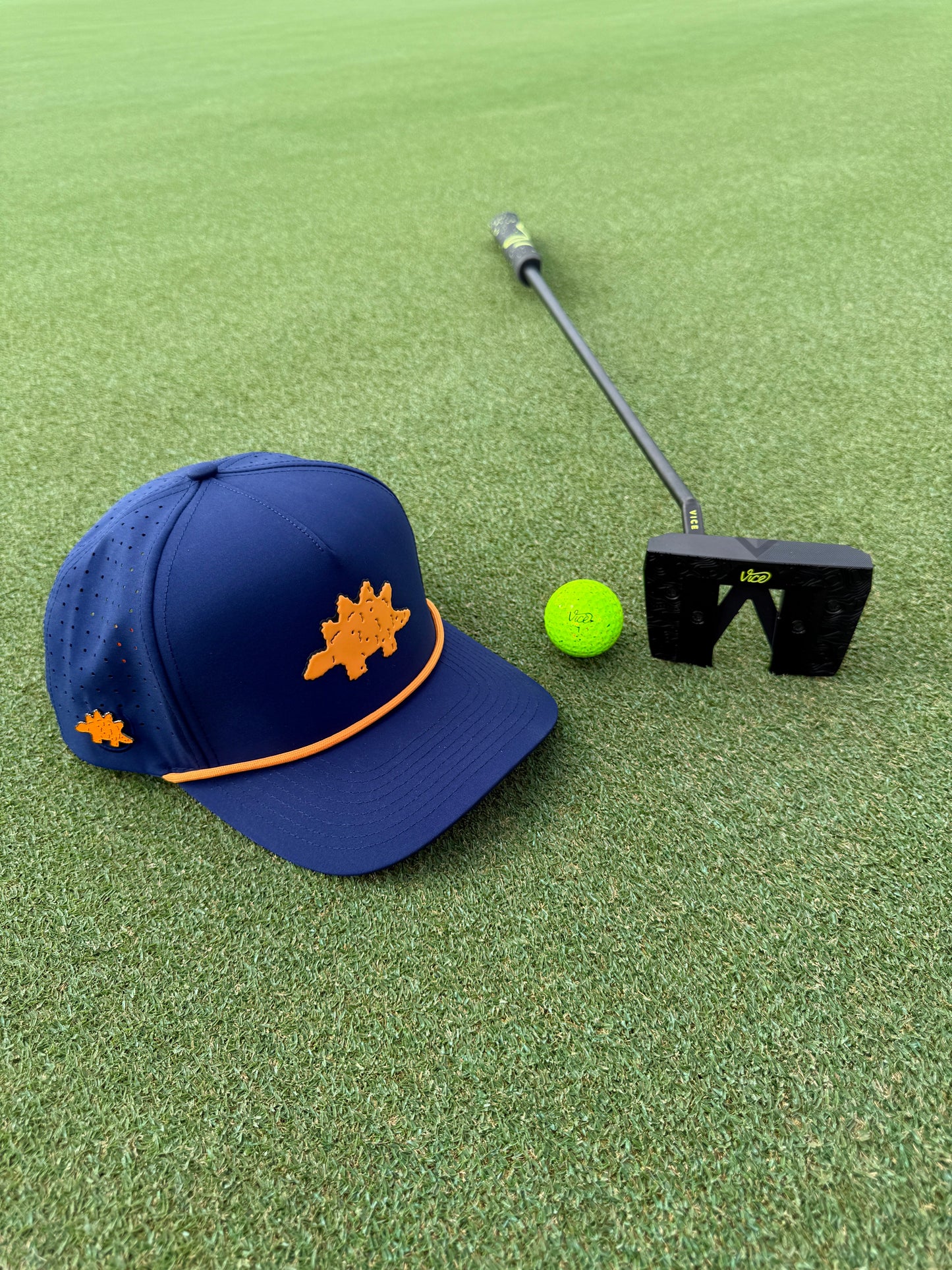 A Logie Joe's Golf Dino Nugget Snapback Hat with a matching ball marker, along with a bright green golf ball and black putter, sit on a well-manicured green.