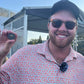 A smiling man in sunglasses, a patterned shirt, and cap holds up The Omen Classic Antique by Doubloon near a tent at East Lake golf course, with trees and blue sky in the background.