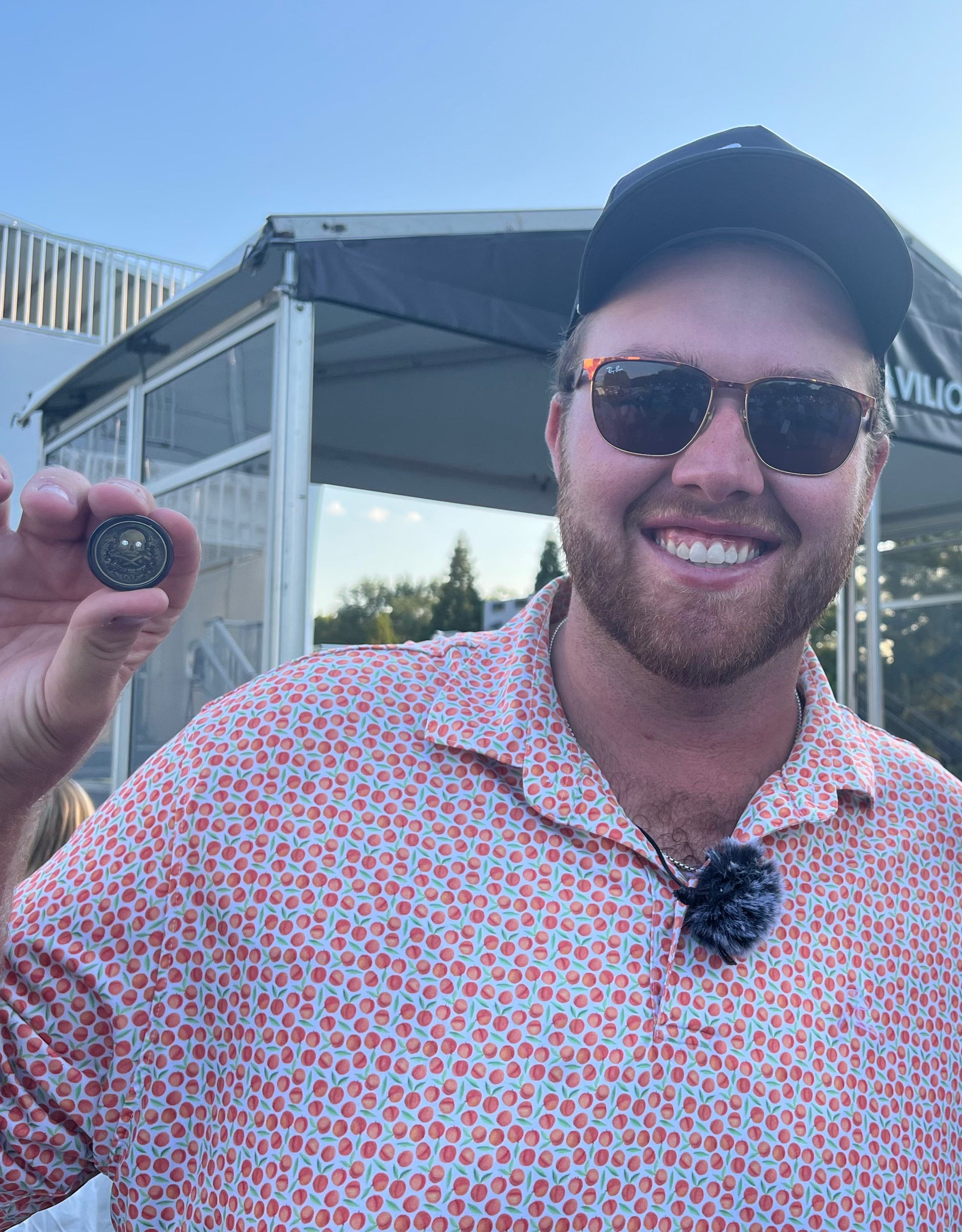 A smiling man in sunglasses, a patterned shirt, and cap holds up The Omen Classic Antique by Doubloon near a tent at East Lake golf course, with trees and blue sky in the background.