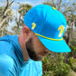 A bearded man wears Logie Joe's Golf Tequila and Lime Snapback Cap with a built-in magnetic ball marker, plus a matching blue shirt, looking downward outdoors among trees and greenery.