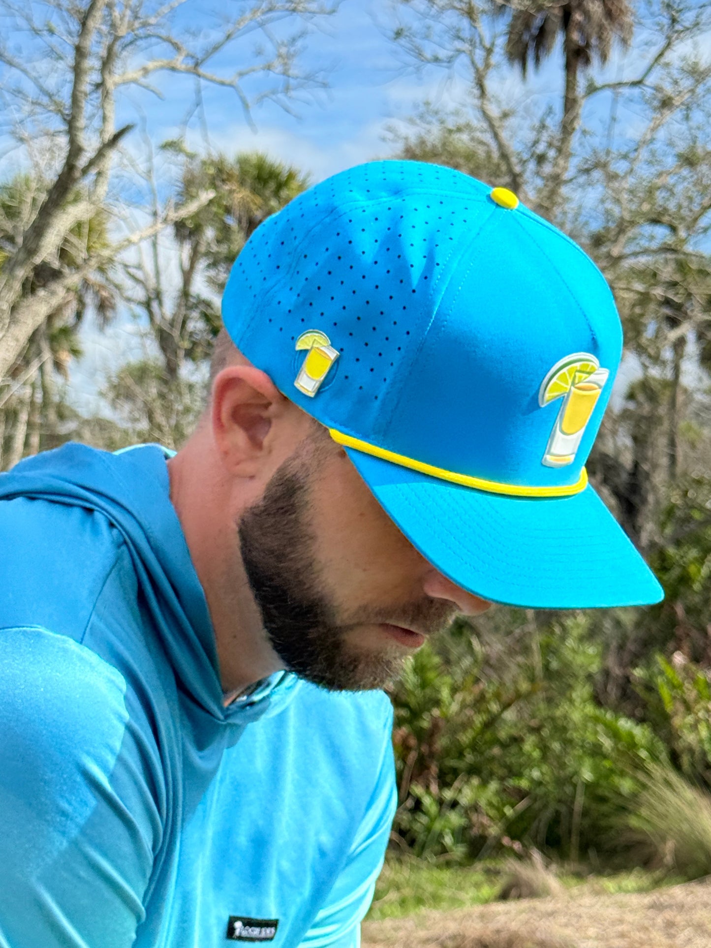 A bearded man wears Logie Joe's Golf Tequila and Lime Snapback Cap with a built-in magnetic ball marker, plus a matching blue shirt, looking downward outdoors among trees and greenery.