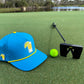 A premium golf accessories setup: Logie Joe's Golf Tequila and Lime Snapback Hat with built-in magnetic ball marker, neon green golf ball, and black putter rest on the green near a water hazard and trees.