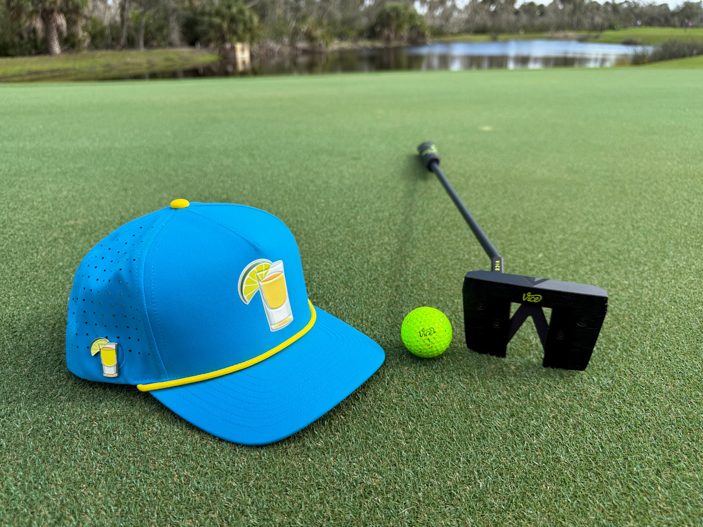 A premium golf accessories setup: Logie Joe's Golf Tequila and Lime Snapback Hat with built-in magnetic ball marker, neon green golf ball, and black putter rest on the green near a water hazard and trees.