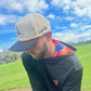 A man wears a gray Logie Joe's Golf Breakfast Ball Snapback Hat with matching ball marker on a green golf course, paired with a black hoodie featuring a colorful hood lining, under a partly cloudy sky.