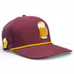 The Logie Joe's Golf Cold One Golf Hat is a maroon adjustable snapback cap with yellow trim, featuring embroidered beer mug designs and a built-in magnetic ball marker holder plus marker, shown on a plain white background.