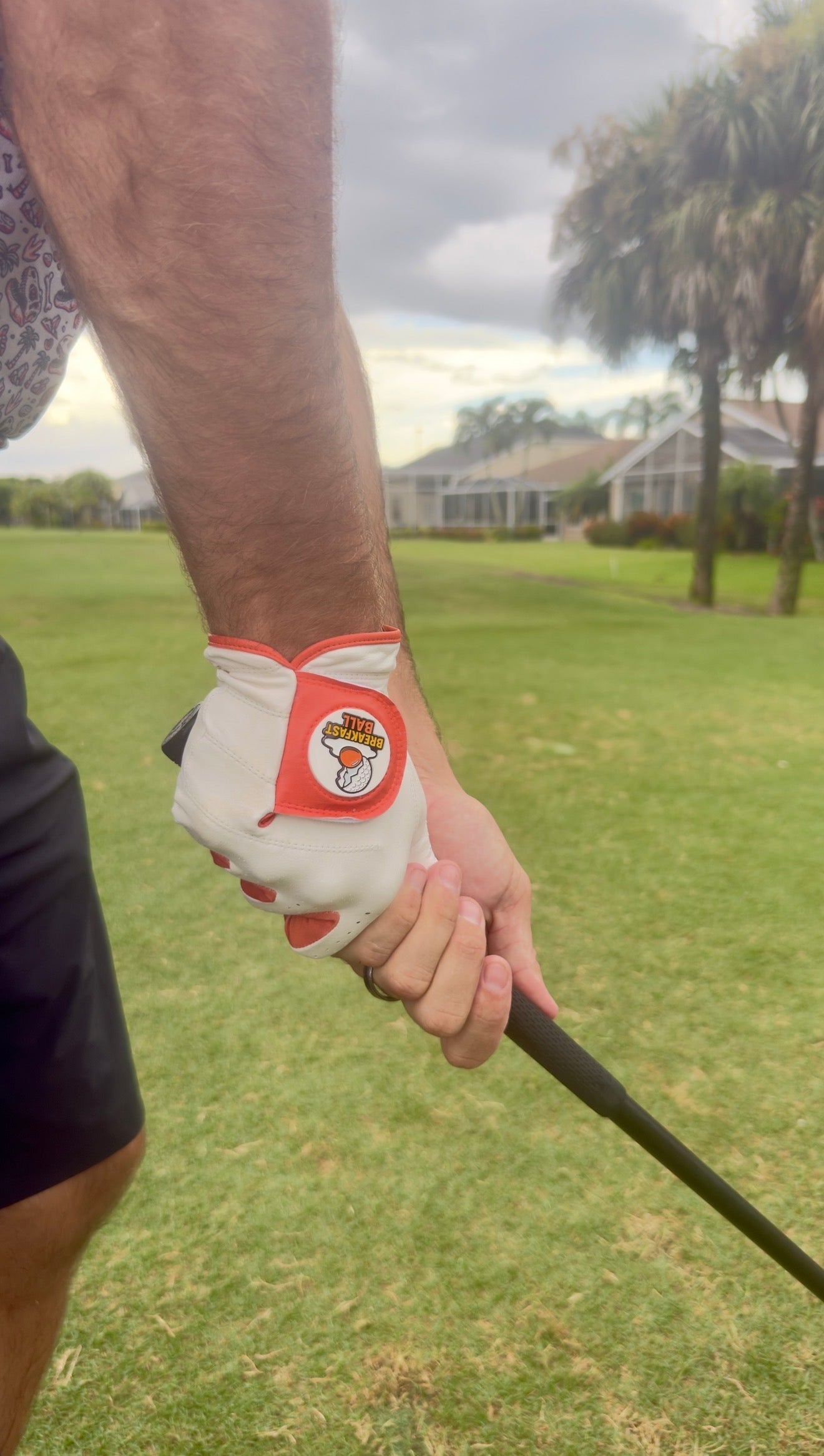 A person wearing the Breakfast Ball Cabretta Leather Golf Glove by Logie Joe’s Golf, a premium glove featuring a magnetic ball marker, grips a golf club on a grassy course with houses and palm trees in the background.