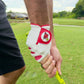 A golfer wearing the Logie Joe’s Golf Transfusion Cabretta Leather Golf Glove—with a built-in magnetic ball marker—grips a yellow club while standing on a green golf course surrounded by houses and trees.