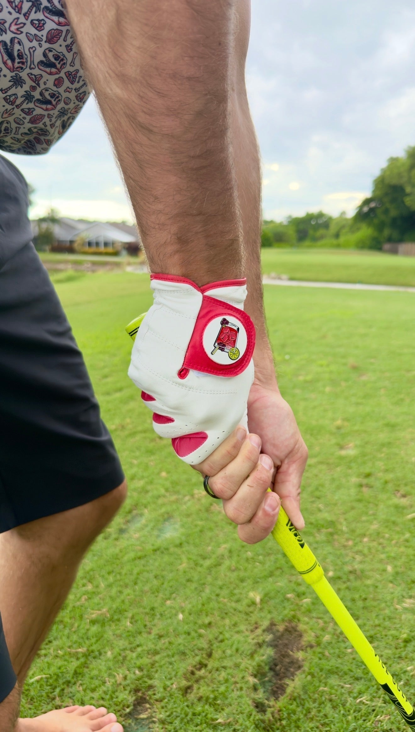 A golfer wearing the Logie Joe’s Golf Transfusion Cabretta Leather Golf Glove—with a built-in magnetic ball marker—grips a yellow club while standing on a green golf course surrounded by houses and trees.