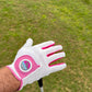 A person wearing a Logie Joe’s Cabretta Leather Golf Glove, featuring premium leather and pink trim, holds out their hand over a grassy area, with a golf club partially visible below.