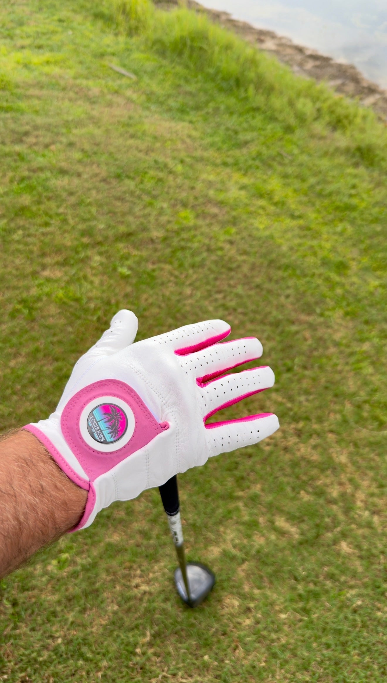 A person wearing a Logie Joe’s Cabretta Leather Golf Glove, featuring premium leather and pink trim, holds out their hand over a grassy area, with a golf club partially visible below.