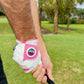 Close-up of a person wearing Logie Joe’s Cabretta Leather Golf Glove, a premium glove featuring a magnetic ball marker, as they grip a golf club on a lush green course with palm trees in the background.