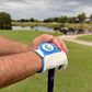 Wearing a Logie Joe’s Golf Water Ball Cabretta Leather Golf Glove—crafted from premium cabretta leather and featuring a convenient magnetic ball marker—a person rests their hands atop a golf club, standing on lush green grass near a scenic pond with trees and cloudy skies in the background.