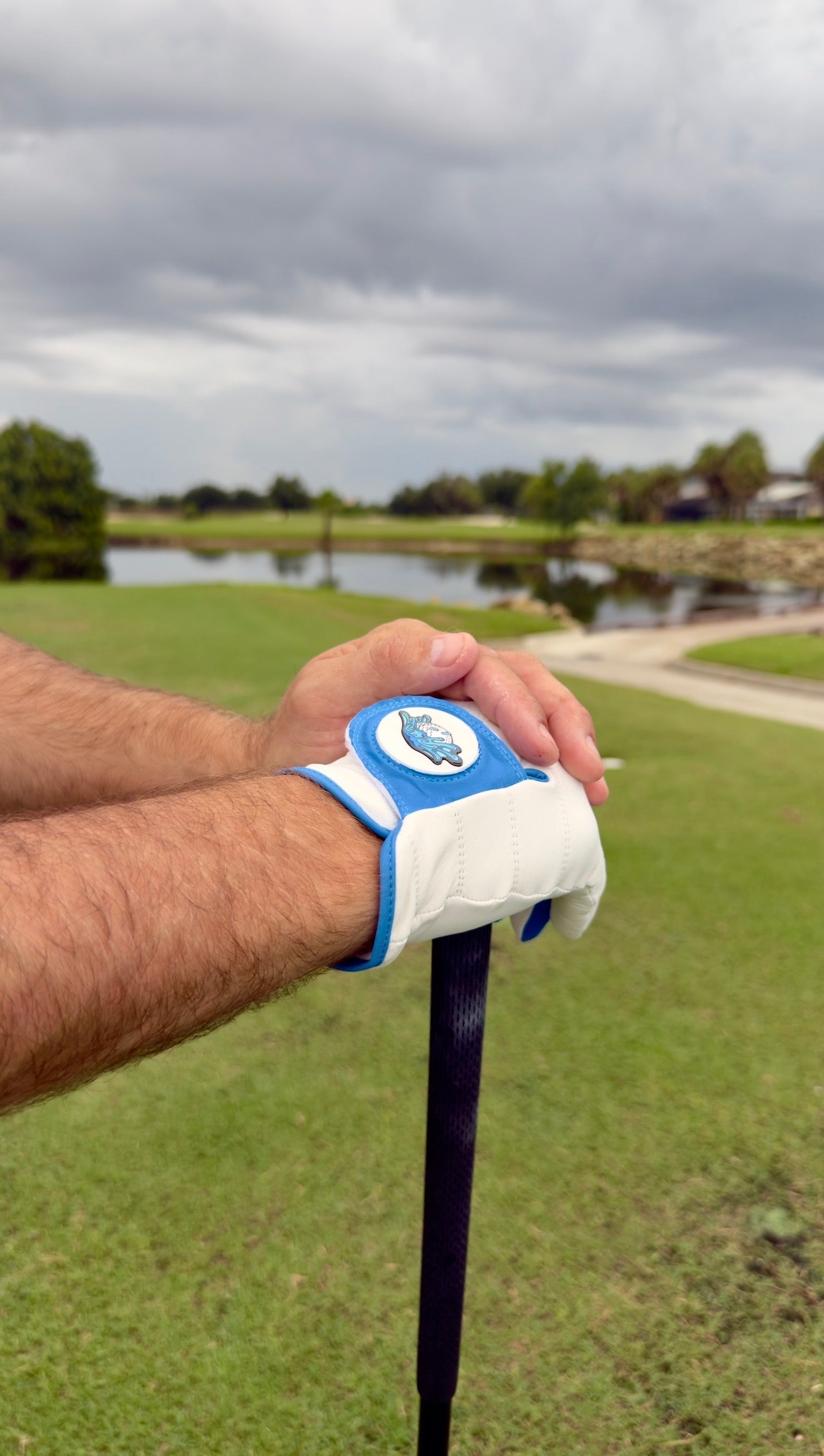Wearing a Logie Joe’s Golf Water Ball Cabretta Leather Golf Glove—crafted from premium cabretta leather and featuring a convenient magnetic ball marker—a person rests their hands atop a golf club, standing on lush green grass near a scenic pond with trees and cloudy skies in the background.