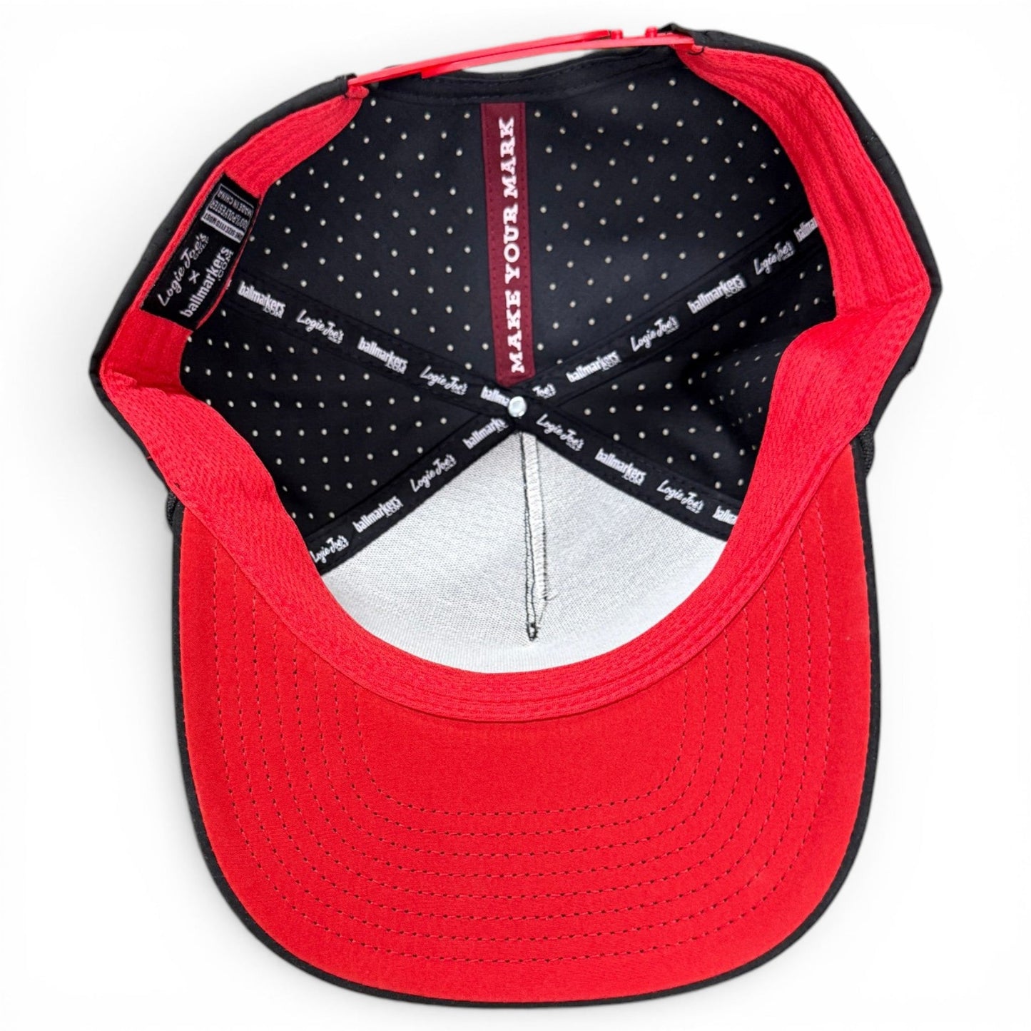 Experience the inside of the Jogie Loe’s Flamingo Golf Hat by Logie Joe’s Golf—a red and black snapback cap featuring a sleek black inner lining patterned with small white dots, signature branded labels, and the inspiring phrase “MAKE YOUR MARK” printed inside the crown. This adjustable golf hat is thoughtfully designed with a built-in magnetic ball marker holder and includes a marker, blending standout style and practical functionality for every golf enthusiast.