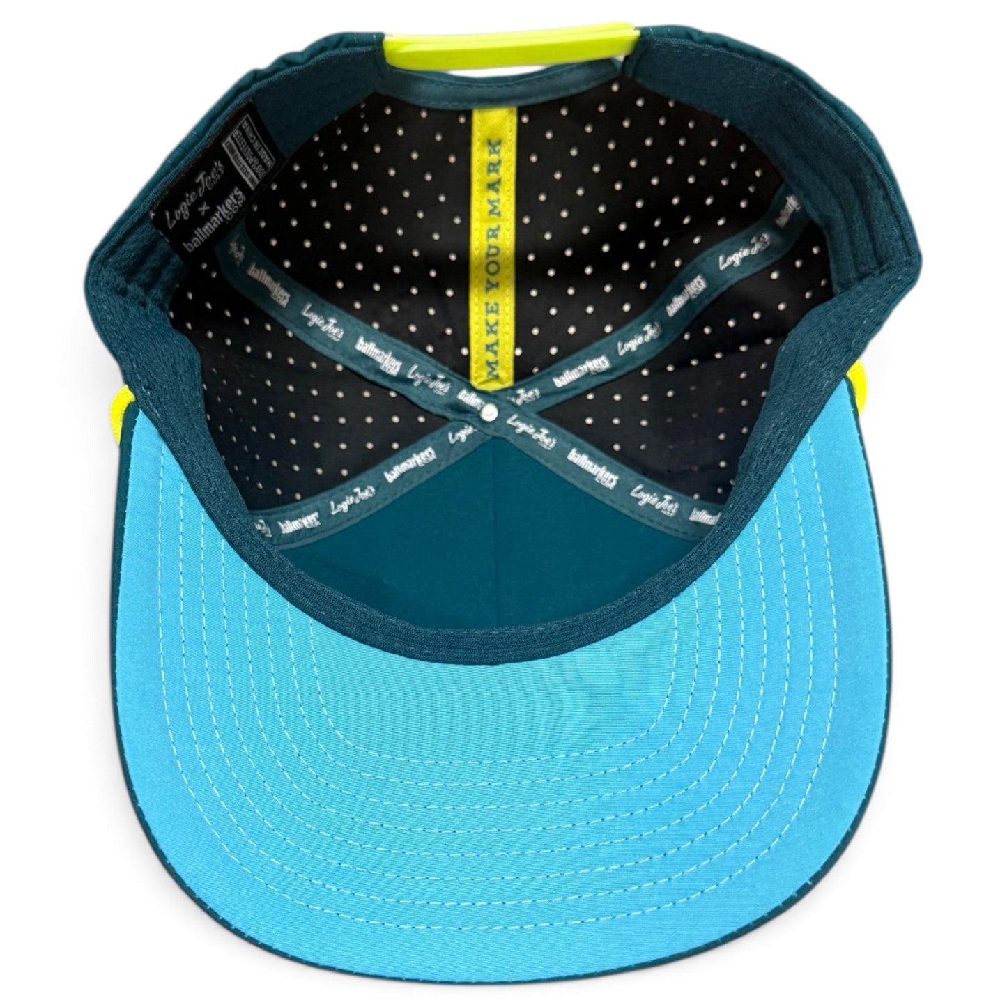 Here’s a rewritten sentence using the provided product data: The inside of the Water Hazard Golf Hat by Logie Joe’s Golf showcases a polka-dot liner, teal sweatband, and a striking yellow band printed with “MAKE YOUR MARK.” This adjustable snapback cap features both blue and black tones and comes equipped with a built-in magnetic ball marker holder—plus marker included—for convenient use on the course.