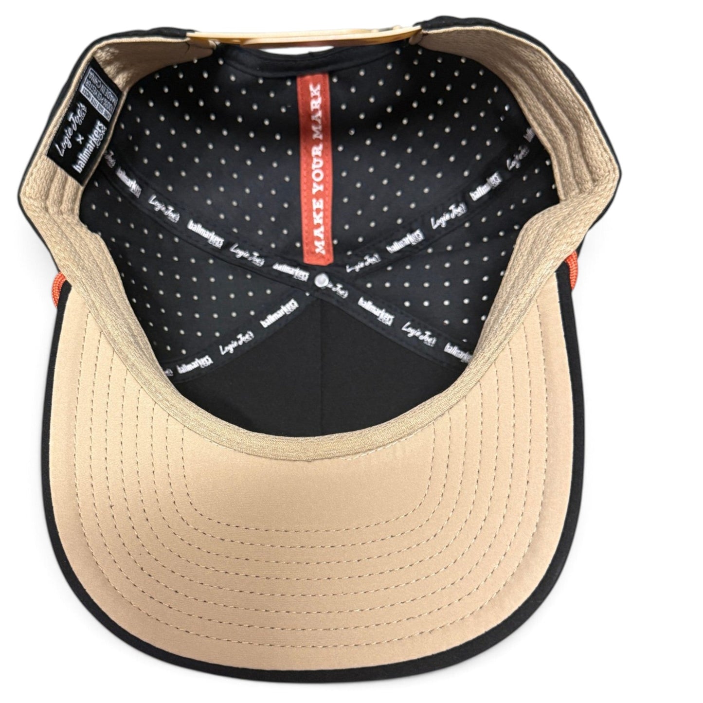 Top view of the inside of the beige and black Logie Joe’s Golf Outlaw Golf Hat, featuring a tan brim, black polka-dot lining, and branded tags and tape along the interior seams. This stylish snapback cap offers an adjustable fit, includes a built-in magnetic ball marker holder with marker, and showcases a distinctive bull skull design.