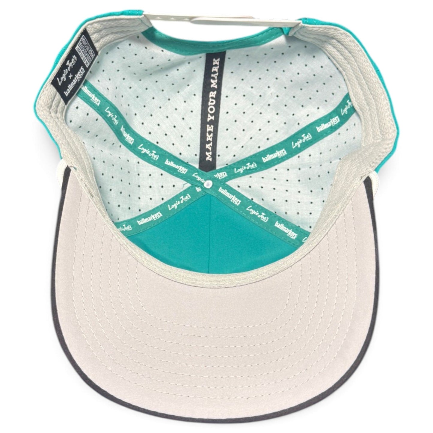 Top-down view of the interior of the Logie Joe’s Golf Mountain Moonrise Golf Hat, featuring a turquoise and beige design. The image highlights the brim, stylish patterned lining, black labels, and a tag reading MAKE YOUR MARK. This adjustable snapback cap also showcases its built-in magnetic ball marker holder with marker included for added convenience.