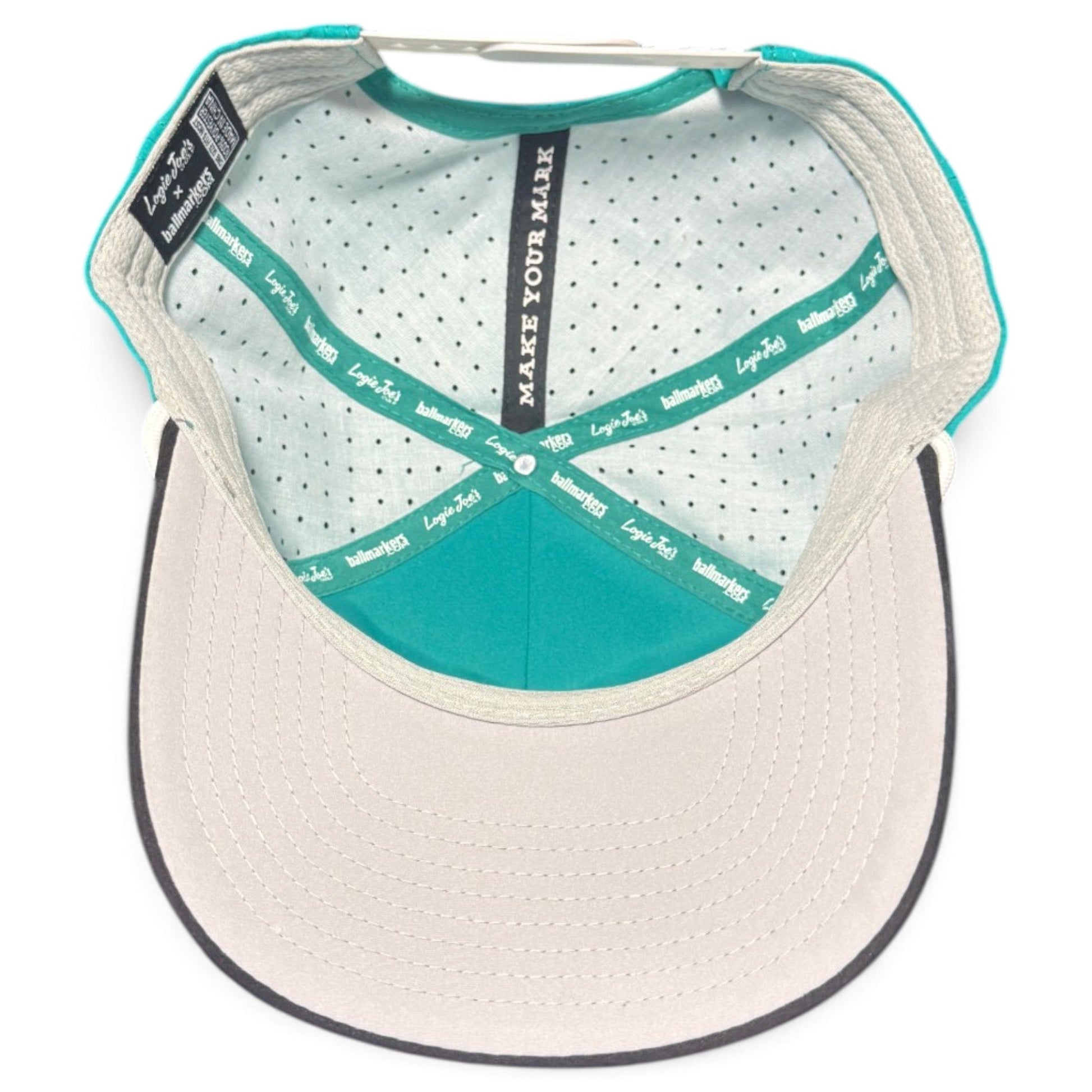 Top-down view of the interior of the Logie Joe’s Golf Mountain Moonrise Golf Hat, featuring a turquoise and beige design. The image highlights the brim, stylish patterned lining, black labels, and a tag reading MAKE YOUR MARK. This adjustable snapback cap also showcases its built-in magnetic ball marker holder with marker included for added convenience.