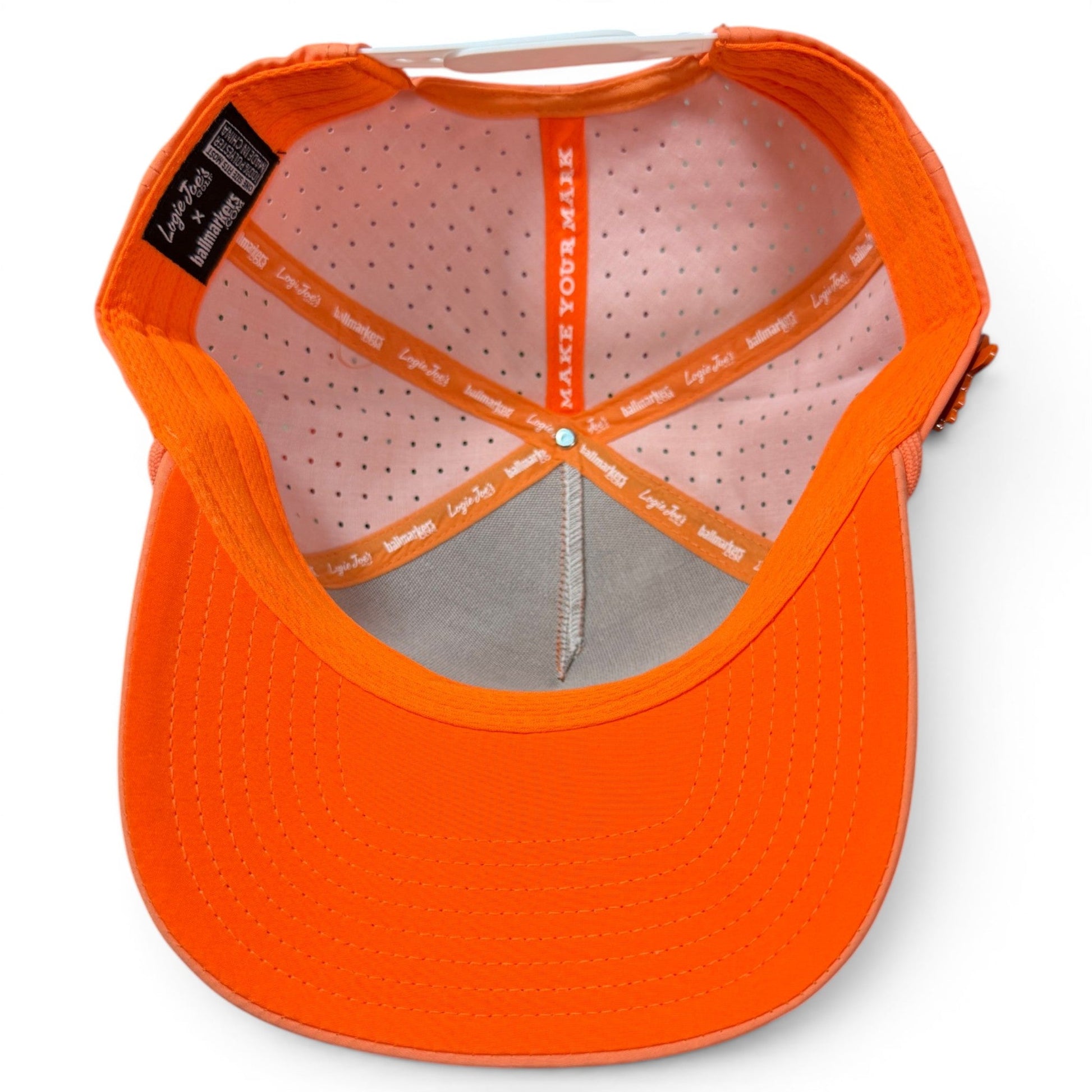 Here is a rewritten version of your sentence using the provided product data: The Logie Joe’s Golf Orange Dream Golf Hat | Snapback Cap is shown from above, revealing its inside lining with white polka dots, an orange sweatband, and interior labels. The phrase MAKE YOUR MARK appears on one of the internal bands, while its built-in magnetic ball marker holder with included marker adds functional appeal—making this adjustable hat both stylish and practical for every round.