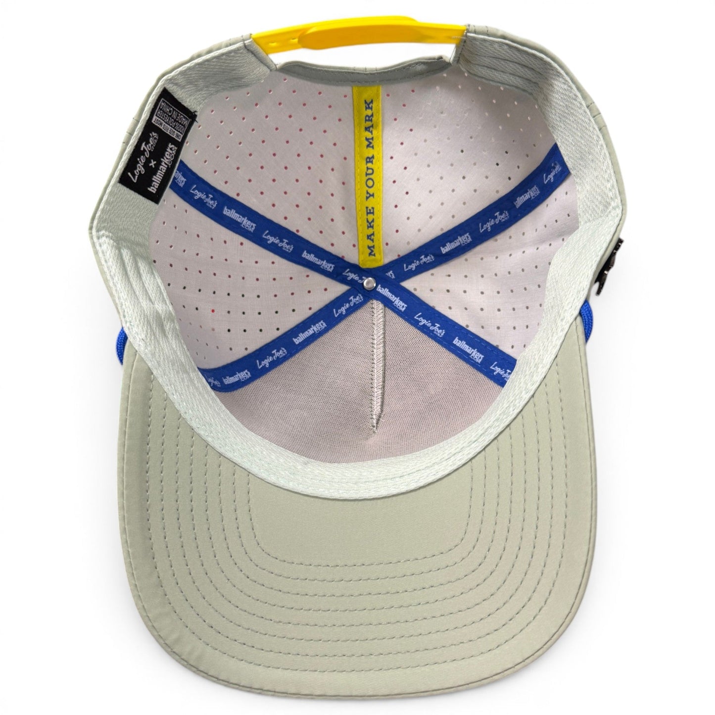 Top view of the inside of a light gray Logie Joe’s Golf Angelfish Golf Hat, featuring blue and yellow accent stitching, a yellow strip with MAKE YOUR MARK text, and interior labels attached to the seams. This stylish snapback cap includes an adjustable fit for customizable comfort and comes equipped with a built-in magnetic ball marker holder plus marker included.