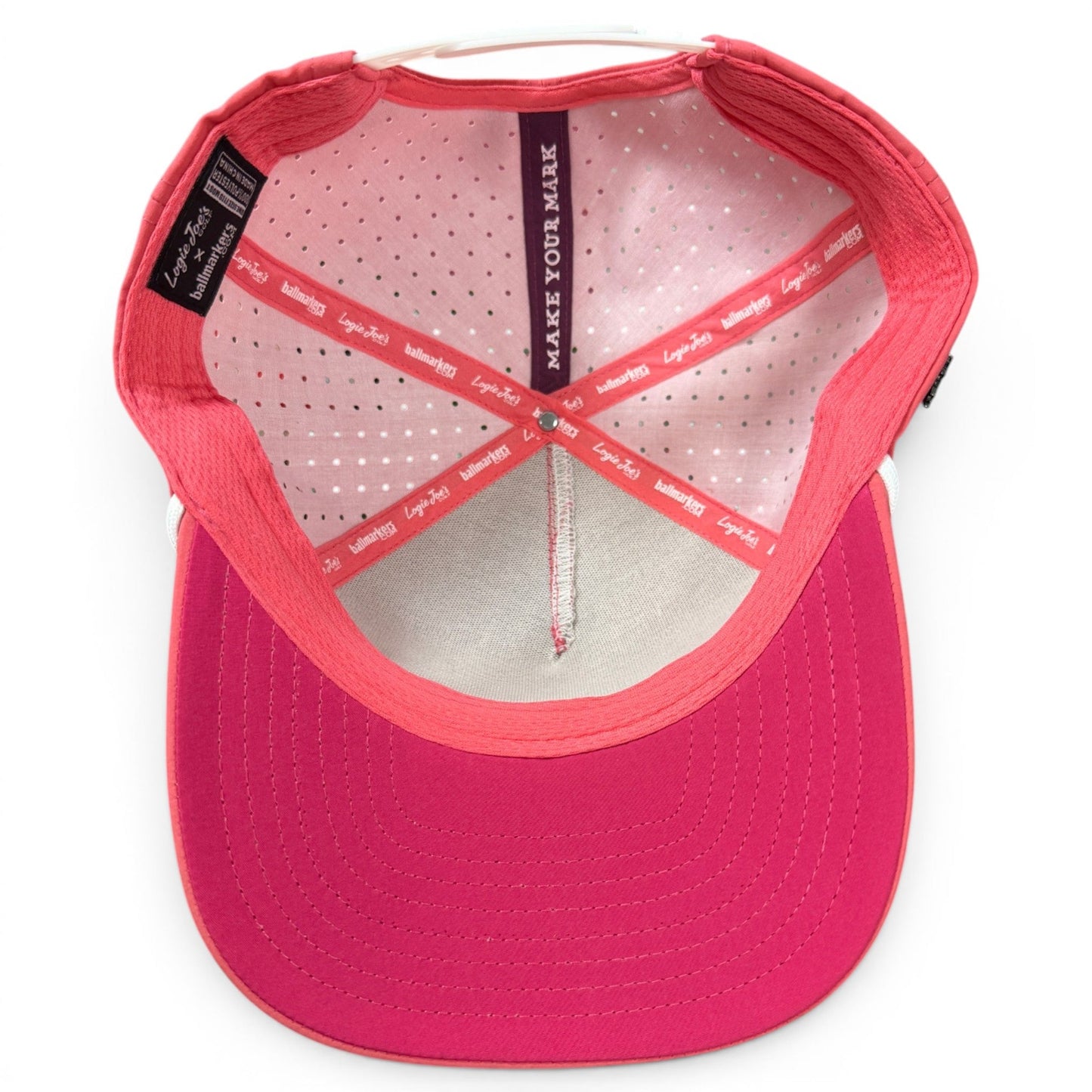 A view of the inside of a pink and white Logie Joe’s Golf Redfoot Birdie Golf Hat | Snapback Cap, featuring stitched panels, a dotted pink lining, and the words MAKE YOUR MARK printed on the inner band—this stylish adjustable red footed booby hat includes a built-in magnetic ball marker holder with marker included.