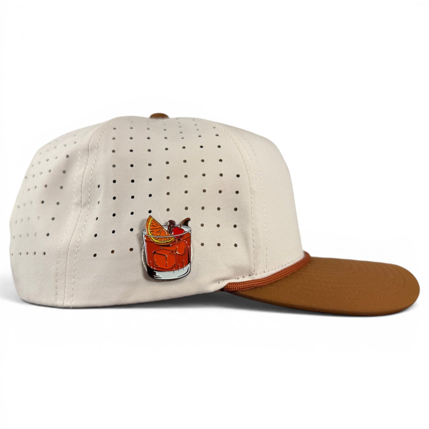 Introducing the Old Fashioned Golf Hat by Logie Joe’s Golf—a cream-colored, adjustable snapback cap designed with small ventilation holes and a brown brim. This stylish golf hat features an enamel pin depicting a cocktail garnished with an orange slice and cherry on the side, plus a built-in magnetic ball marker holder with included marker for added convenience on the course.