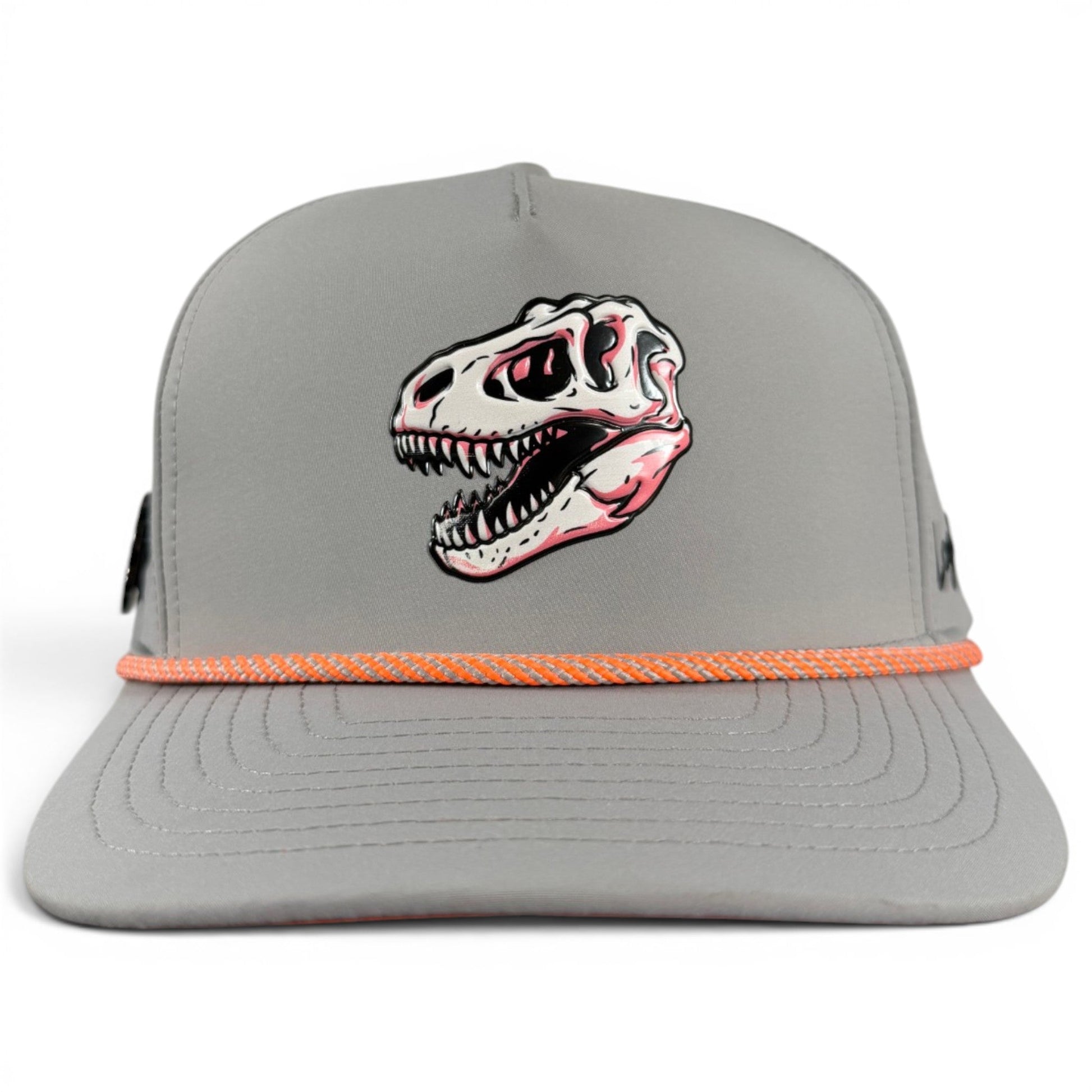 Introducing the Fossil Fairway Golf Hat by Logie Joe’s Golf—a gray snapback cap featuring an illustrated dinosaur skull on the front and a striking orange rope detail above the brim. This adjustable golf hat seamlessly blends style and function with its built-in magnetic ball marker holder (marker included), making it the perfect accessory for keeping your game on point.