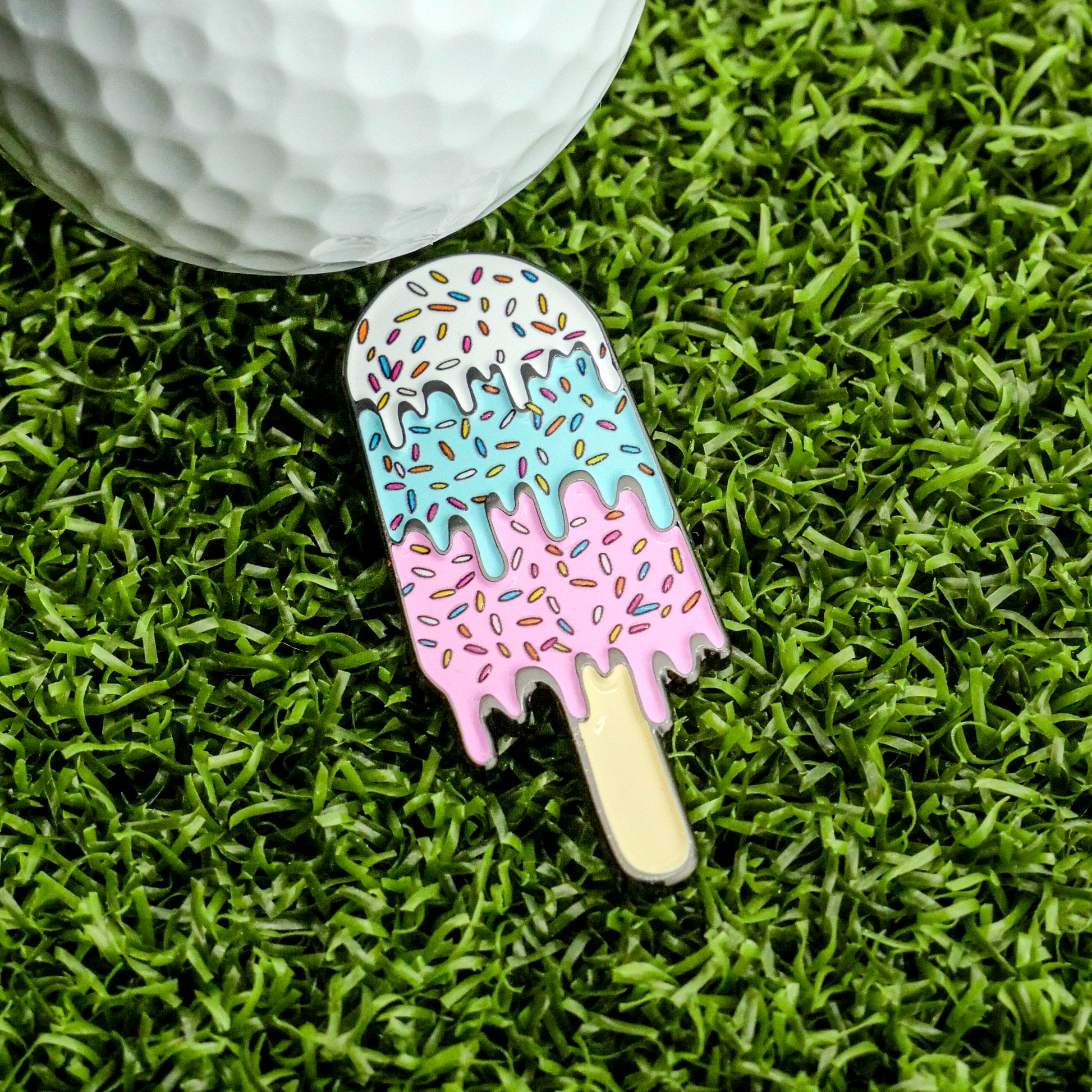 The Ice Cream Bar Ball Marker x Shank-It Golf from Ballmarkers.com features a colorful enamel design shaped like a melting ice cream popsicle with sprinkles, shown next to a white golf ball on green artificial grass.