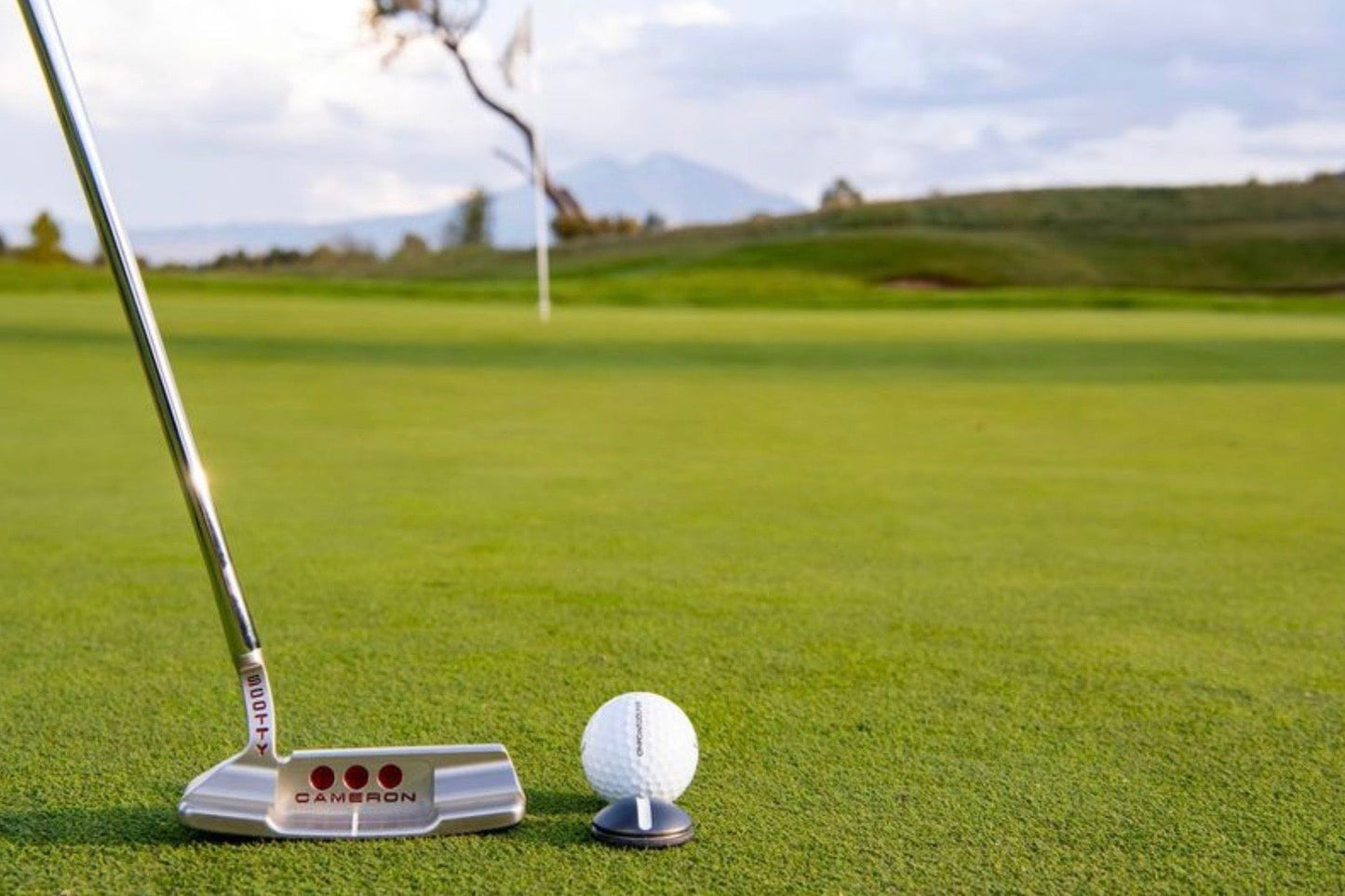 A close-up of an OnPointGolf.us One Rail Smooth Ball Marker used as a putting aid with a golf putter and ball on green grass; a flag and trees appear blurred in the background.