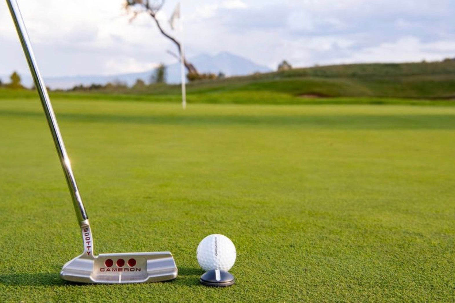 A close-up of an OnPointGolf.us One Rail Smooth Ball Marker used as a putting aid with a golf putter and ball on green grass; a flag and trees appear blurred in the background.