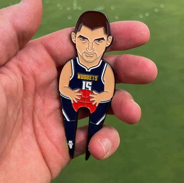 A hand holds Pin Creatures' Joker, a novelty divot tool shaped like a basketball player in a Nuggets #15 jersey holding a ball, with an attached golf ball marker, set against a green blurred background.