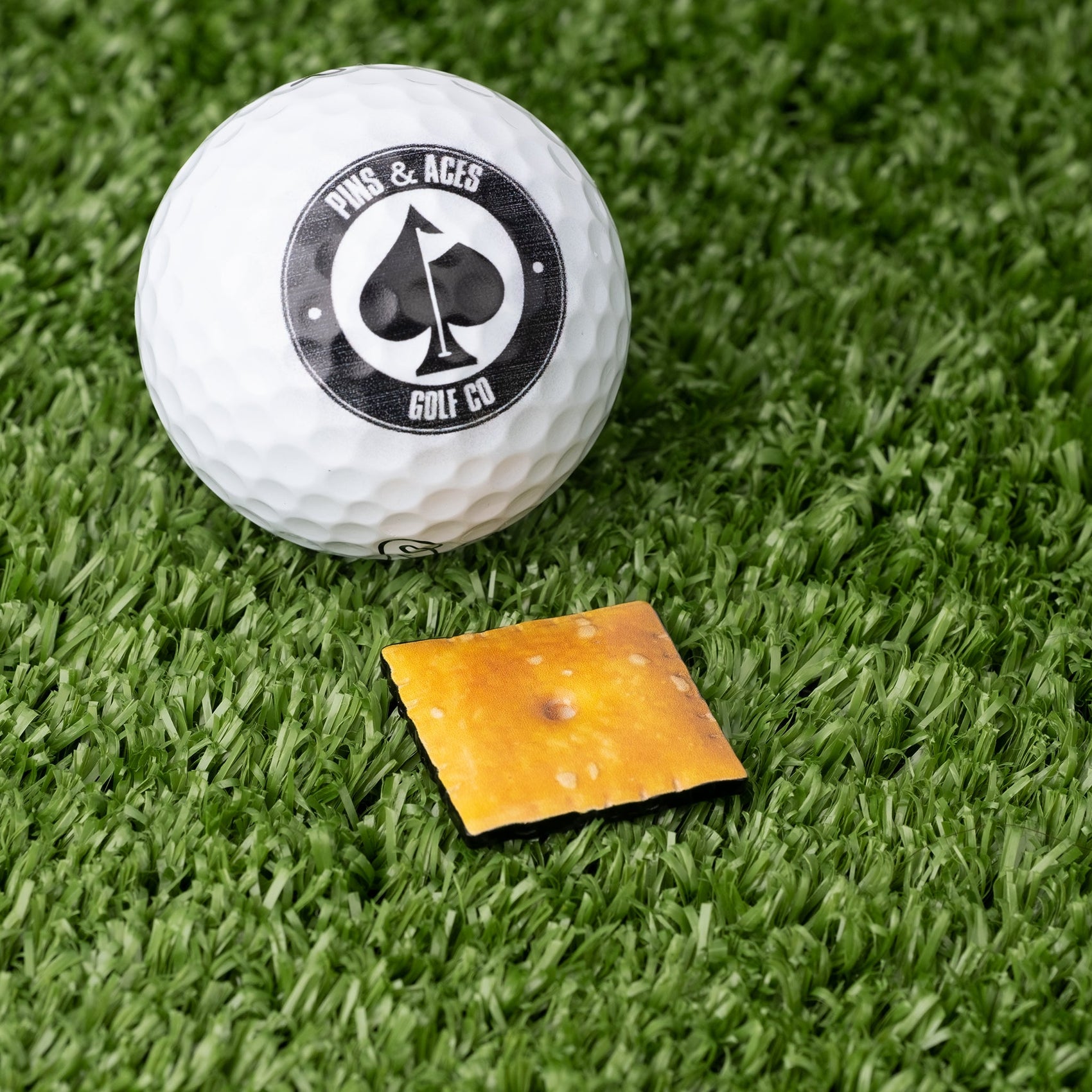 A white golf ball with a Pins and Aces logo sits on green turf next to a small square Cheese Cracker Ball Marker from Pins and Aces.