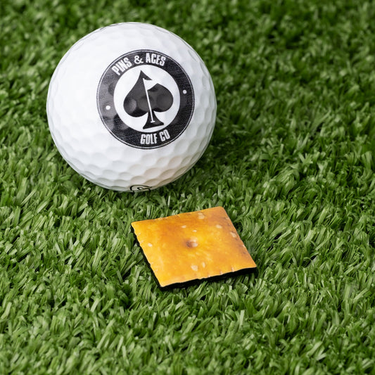 A white golf ball with a Pins and Aces logo sits on green turf next to a small square Cheese Cracker Ball Marker from Pins and Aces.