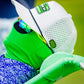 A person in a bright green face cover, sunglasses, white cap with green/black accents, and a blue patterned shirt holds light green equipment and a Pins and Aces HEYDUDE Logo Ball Marker, possibly on a golf course.