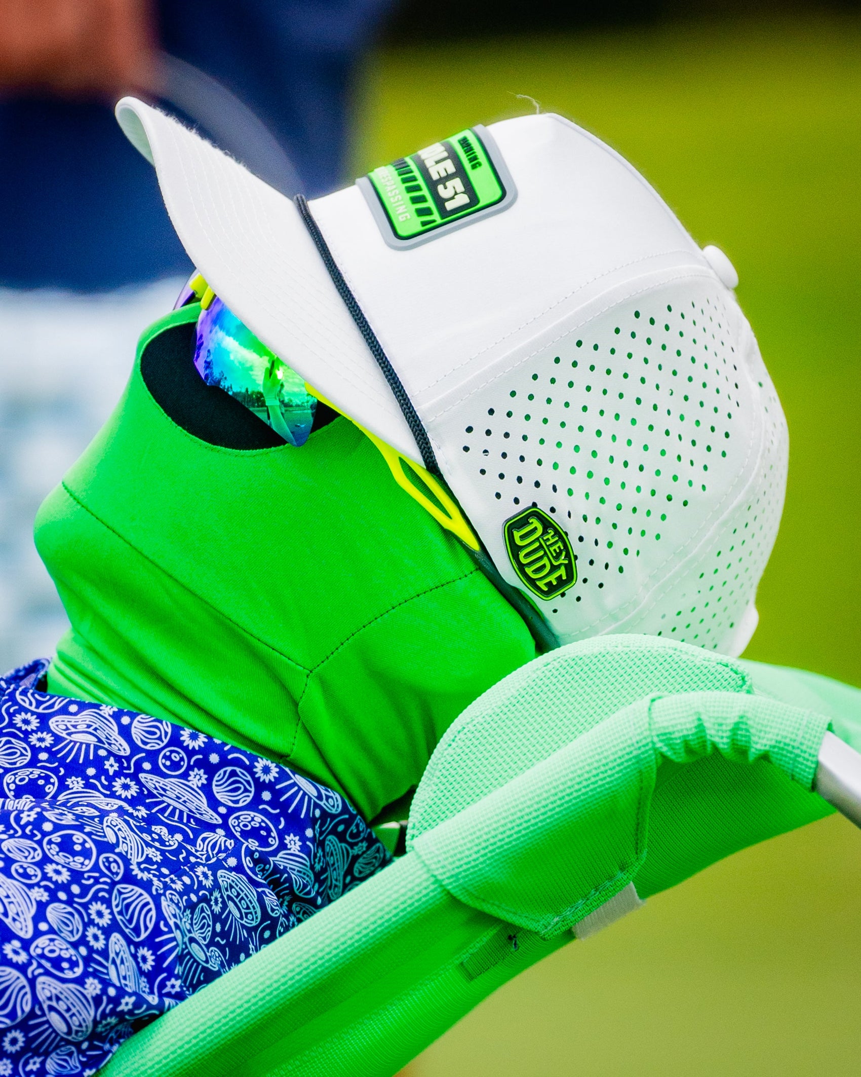 A person in a bright green face cover, sunglasses, white cap with green/black accents, and a blue patterned shirt holds light green equipment and a Pins and Aces HEYDUDE Logo Ball Marker, possibly on a golf course.