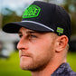 A bearded man wears a black HEYDUDE cap, a patterned shirt, and a Pins and Aces Hole 51 Ball Marker clipped to his hat. He stands outdoors amid blurred greenery, looking to the left with a neutral expression.