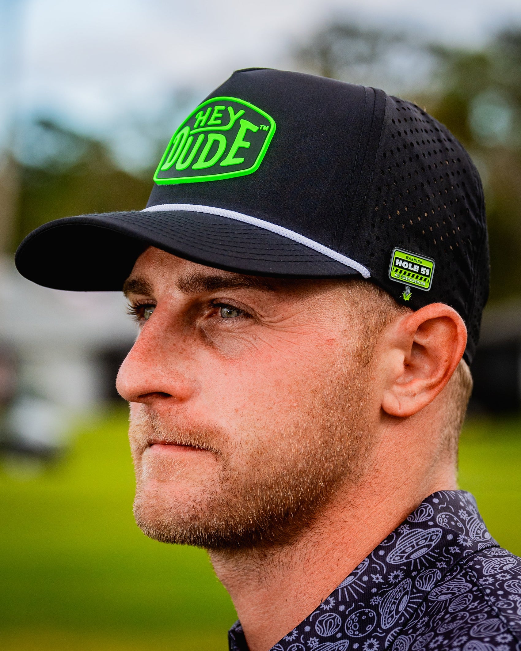 A bearded man wears a black HEYDUDE cap, a patterned shirt, and a Pins and Aces Hole 51 Ball Marker clipped to his hat. He stands outdoors amid blurred greenery, looking to the left with a neutral expression.