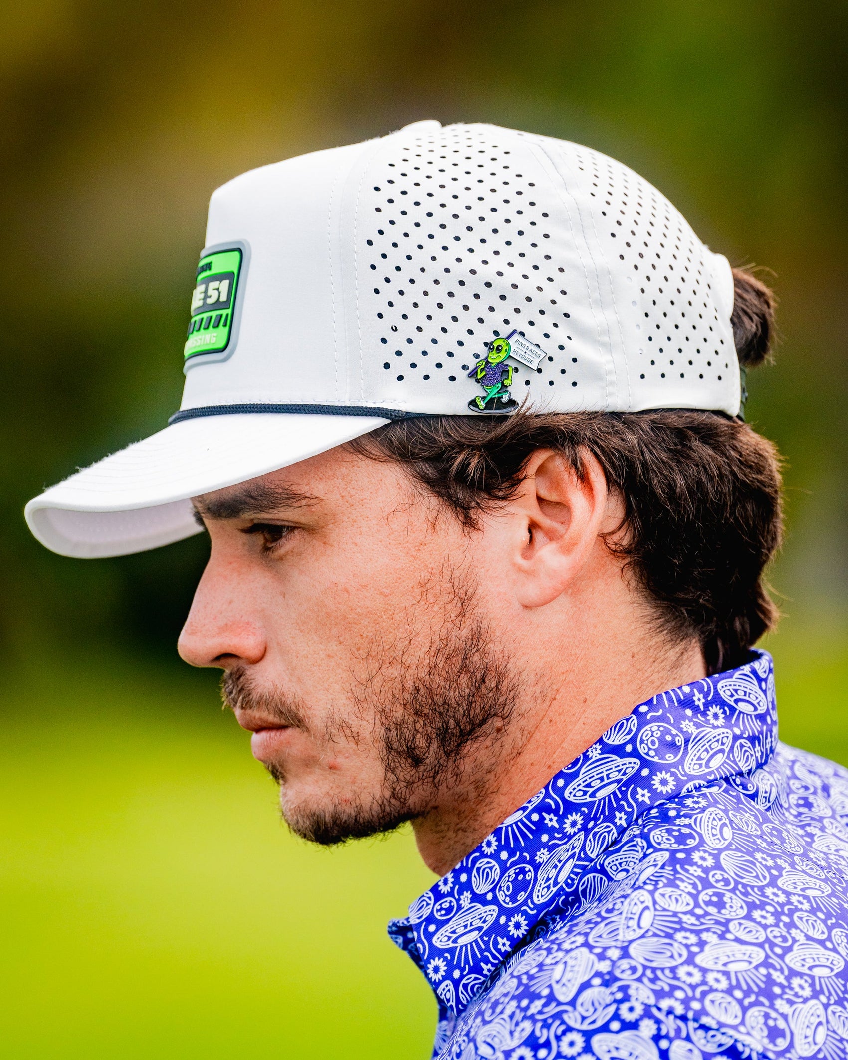 A man with short dark hair and a beard stands outdoors in a white perforated cap featuring a golfer graphic, wearing a blue patterned collared shirt. His cap is adorned with the Pins and Aces HEYDUDE - Alien Ball Marker.